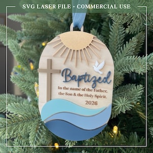 Puede incluir: Adorno de bautizo de madera con un dise&ntilde;o de cruz, paloma y sol. El adorno tiene la palabra "Baptized" y el texto "In the name of the Father, the Son & the Holy Spirit. 2026". Colgando de un &aacute;rbol de Navidad.