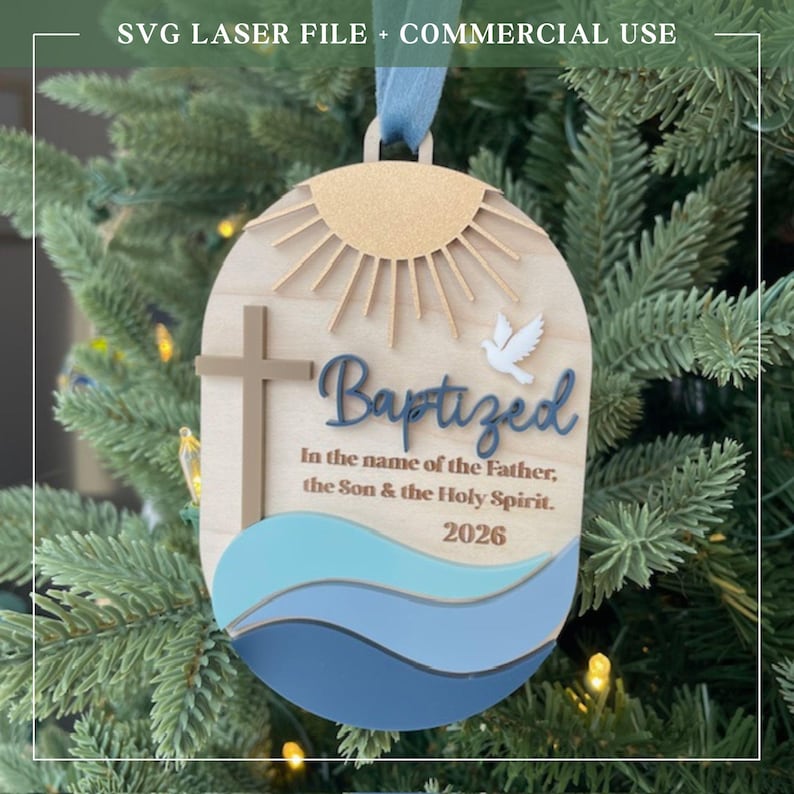 Puede incluir: Adorno de bautizo de madera con un dise&ntilde;o de cruz, paloma y sol. El adorno tiene la palabra "Baptized" y el texto "In the name of the Father, the Son & the Holy Spirit. 2026". Colgando de un &aacute;rbol de Navidad.