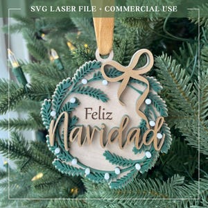 May include: A round wooden Christmas ornament featuring a laser-cut design of green pine branches and small white berries. The text Feliz Navidad is engraved and layered in the center. A gold ribbon is attached to the top for hanging purposes.