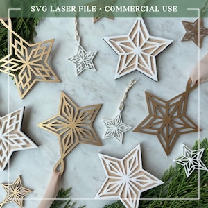 Pode incluir: Uma coleção de ornamentos em forma de estrela em tons de dourado, branco e madeira natural, com recortes geométricos intrincados. Algumas estrelas incluem fitas ou cordões para pendurar. O texto SVG LASER FILE + COMMERCIAL USE é visível.