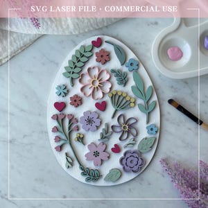 May include: A white wooden egg-shaped ornament decorated with laser-cut floral designs in shades of pink, green, and purple. The ornament is surrounded by a white background.