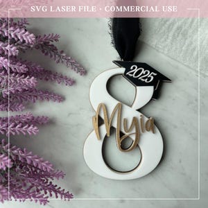 Könnte beinhalten: Weißer Holzschmuck in Form der Zahl Acht mit einer schwarzen Abschluss-Kappe oben drauf. Die Kappe hat das Jahr 2025 darauf gedruckt. Der Name "Myia" ist in goldenen Glitzer auf dem Ornament geschrieben.