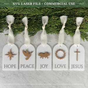 May include: Five white arched ornaments with cream ribbons, each featuring a gold-toned icon and text: a star labelled HOPE, a dove labelled PEACE, a manger labelled JOY, a crown of thorns labelled LOVE, and a cross labelled JESUS.