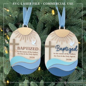 Puede incluir: Dos adornos de bautizo de madera con cintas azules. Cada adorno presenta una cruz, una paloma y la palabra "Baptized" con el texto "In the name of the Father, the Son & the Holy Spirit. 2026". La parte superior tiene un dise&ntilde;o de sol.