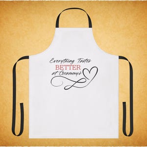 以下が含まれることがあります： 黒いストラップが付いた白いエプロン。「Everything Tastes BETTER at Grammy's」というテキストが黒と赤のスクリプトで書かれています。ハートと装飾のデザイン。キッチンでの使用に最適です。