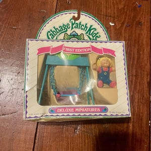 May include: A vintage Cabbage Patch Kids Deluxe Miniatures set, First Edition. The box features a teal swing set and a doll in overalls. The packaging includes the text "Cabbage Patch Kids", "First Edition", and "Deluxe Miniatures".