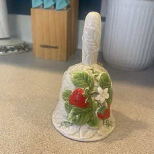 May include: A ceramic bell with a white base and handle, decorated with red strawberries, green leaves, and white flowers. The bell has a detailed, embossed design and a small gold-colored accent near the top.