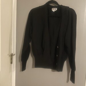 May include: A black cardigan sweater with long sleeves and a V-neck. The sweater has three silver buttons down the front and is hanging on a black hanger. The label reads "Adele Joyce".