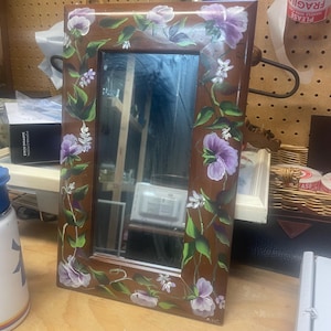 May include: A rectangular brown framed mirror with hand-painted floral designs. The frame is adorned with purple and white flowers, green leaves, and vines. The mirror reflects the surrounding environment.