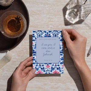 Peut inclure: Une carte de vœux avec un motif floral bleu et le texte "Je te vois, je me soucie de toi, et Jéhovah aussi." La carte est tenue par deux mains. La carte a un centre bleu clair avec le texte. L'arrière-plan comprend une boisson et un objet décoratif.