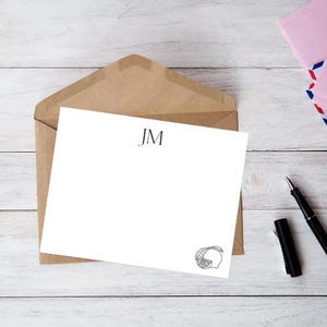 Puede incluir: Papelería blanca con las iniciales "JM" y una ilustración de limón, sobre un sobre marrón. Un bolígrafo negro y un sobre rosa con rayas rojas y azules también son visibles.