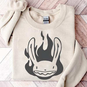 Può includere: Felpa beige con grafica grigio scuro di una testa di coniglio in fiamme. La felpa ha girocollo e maniche lunghe con polsini a coste. L'etichetta del marchio Gildan è visibile all'interno del colletto.