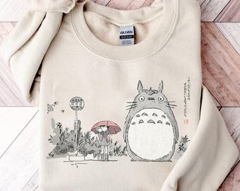 May include: Beige crewneck sweatshirt featuring a graphic of a large, gray cartoon character with a white belly, a girl with a red umbrella, and a bus stop. The design is in a minimalist style with Japanese text.