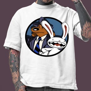 Può includere: T-shirt bianca con una grafica a cartoni animati di Sam & Max, un duo cane e coniglio, all'interno di un cerchio nero. Il cane indossa un abito e una cravatta, e il coniglio è bianco con lunghe orecchie. La maglietta è indossata da una persona con tatuaggi e piercing.