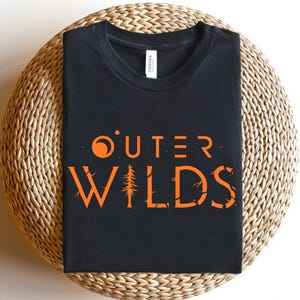 Op de afbeelding: Zwart t-shirt met de woorden "OUTER WILDS" in oranje, met een gestileerde zon en boomafbeelding. Het shirt wordt getoond op een geweven, cirkelvormig oppervlak.