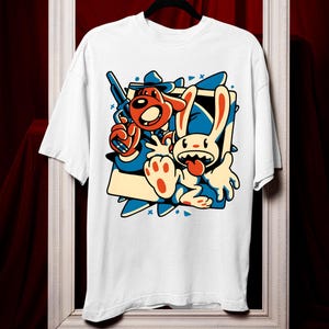 Può includere: T-shirt bianca con un'illustrazione a cartoni animati di un cane che tiene una pistola e un coniglio con la lingua di fuori. Il design utilizza colori rosso, blu e giallo. La maglietta è appesa a una gruccia nera su uno sfondo rosso.