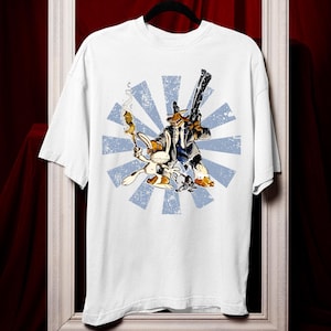 Può includere: T-shirt bianca con un'illustrazione a cartoni animati di un coniglio e un cane in giacca e cravatta, che tengono una pistola e un sigaro acceso. La grafica è ambientata su un design a raggi di sole blu.