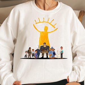 Può includere: Felpa bianca con grafica gialla di una figura con un design a occhio sopra un gruppo di personaggi dei cartoni animati. I personaggi includono un orso in giacca e cravatta, un coniglio e altre figure. La felpa è girocollo.