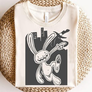 Può includere: T-shirt color crema con un'illustrazione a fumetti in bianco e nero di un personaggio coniglio che tiene una spada, con uno skyline sullo sfondo. Il coniglio ha un sorriso malizioso ed è in movimento. La maglietta è esposta su una superficie tessuta.