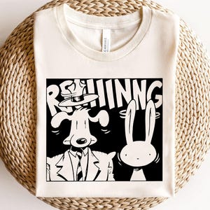 Può includere: T-shirt color crema con grafica in bianco e nero di un cane in giacca e cravatta e un coniglio, con la scritta "REHINNG". La maglietta è realizzata in materiale morbido ed è esposta su una superficie tessuta.