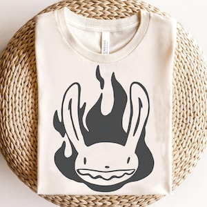 Può includere: T-shirt color crema con grafica nera di un volto di coniglio in fiamme. Il coniglio ha lunghe orecchie e un ampio sorriso. La maglietta è su una superficie circolare tessuta.
