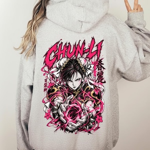 Puede incluir: Una sudadera con capucha de color gris claro que presenta un gran gráfico en la parte posterior. El diseño muestra al personaje Chun-Li con cabello oscuro, rodeado de remolinos de energía rosa y flores de cerezo, con el texto CHUN-LI.