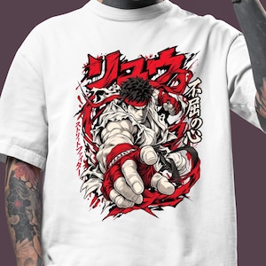 Op de afbeelding: Een wit T-shirt met korte mouwen en een grafische illustratie van het personage Ryu uit Street Fighter. Het ontwerp bevat rode accenten, Japanse tekens en een dynamische pose met een vuist die naar voren is gericht.