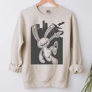 Può includere: Felpa beige con un personaggio di coniglio dei cartoni animati e uno sfondo di paesaggio urbano. Il coniglio è raffigurato in una posa dinamica, che indica con una mano e tiene in mano un'arma. Il design è in bianco e nero, creando un contrasto con il tessuto colorato neutro.