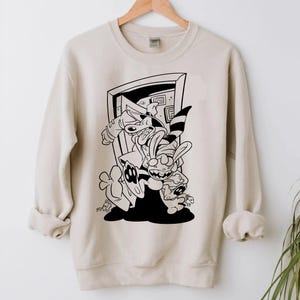 Può includere: Felpa beige con grafica a fumetti in bianco e nero. La grafica raffigura un personaggio simile a un cane e un personaggio simile a un coniglio davanti a una porta. La felpa ha maniche lunghe e girocollo.