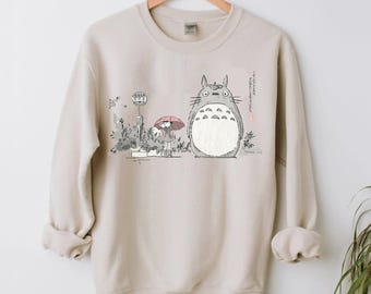 May include: Beige sweatshirt featuring a graphic of Totoro from My Neighbor Totoro. The illustration shows Totoro with a girl holding an umbrella at a bus stop. The design includes Japanese text and a bus stop sign.