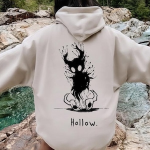 Op de afbeelding: Beige hoodie met een zwarte afbeelding op de rug met een gestileerd wezen met een donker, gespat ontwerp en het woord "Hollow". De hoodie heeft een capuchon en lange mouwen.