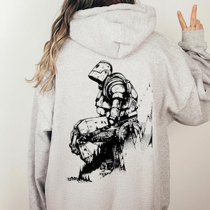 May include: A light grey hoodie featuring a black and white illustration of a robot on the back. The robot is depicted in a seated position, with intricate detailing. The hoodie has a hood and long sleeves.