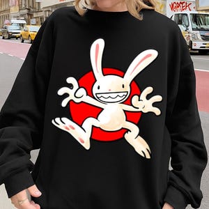 Può includere: Felpa nera con un design a personaggio di coniglio dei cartoni animati. Il coniglio è bianco con lunghe orecchie e un ampio sorriso, su un cerchio rosso. Il design è centrato sulla parte anteriore della felpa.