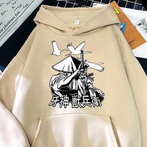May include: Beige hoodie with a front pocket and a hood. The front features a black and white graphic of a samurai figure with a sword, a conical hat, and two flying birds. Japanese text is below the graphic.