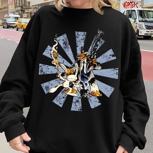 Può includere: Felpa nera con grafica a cartoni animati di un coniglio e un cane con pistole, su un design a raggi di sole blu. La felpa è indossata con un berretto da baseball beige. Lo sfondo mostra una strada cittadina con edifici e veicoli.