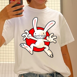 Può includere: T-shirt bianca con un disegno di coniglio dei cartoni animati su uno sfondo a cerchio rosso. Il coniglio ha un ampio sorriso ed è in una posa dinamica. Un capo casual per tutti i giorni.