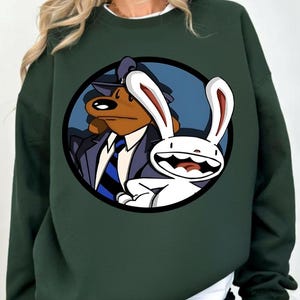 Può includere: Felpa verde scuro con una grafica circolare di Sam & Max, un duo di cani e conigli dei cartoni animati. Il cane indossa un abito e un cappello, e il coniglio è bianco con grandi orecchie. La grafica è su sfondo blu.