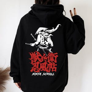 May include: Black hooded sweatshirt featuring a white and red graphic of a samurai warrior with swords and Japanese text. The text reads "NINJA SCROLL" below the illustration. The design is on the back of the hoodie.