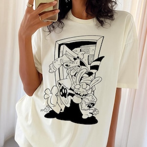 Può includere: T-shirt color crema con grafica a fumetti in bianco e nero di due personaggi in una porta. La grafica include un coniglio con un cappello a righe e un personaggio simile a un cane. La maglietta ha maniche corte e una vestibilità ampia.