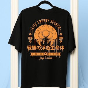 Puede incluir: Camiseta negra con un diseño gráfico naranja. El diseño incluye el texto "LIFE ENERGY SEEKER" y caracteres japoneses. El gráfico también presenta una criatura estilizada y las palabras "FLOATING LIFE FORM" y "Stage 3, larva."