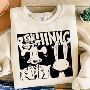 Può includere: Felpa color crema con grafica in bianco e nero di un cane in giacca e cravatta e un coniglio, con la scritta "REHINNG" sopra. La felpa ha girocollo e maniche lunghe.