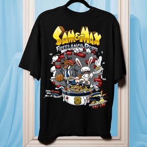 Può includere: T-shirt nera con una grafica colorata di Sam & Max: Freelance Police. Il design include il testo "Sam & Max Freelance Pops" e un'illustrazione a cartoni animati dei personaggi con una ciotola di cereali. La maglietta è esposta su una gruccia.