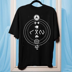 Op de afbeelding: Zwart T-shirt met een wit cirkelvormig ontwerp met geometrische vormen en symbolen. Het ontwerp bevat een driehoek, een gestileerde figuur en andere abstracte elementen. Het shirt wordt op een hanger gepresenteerd tegen een lichtblauwe achtergrond.