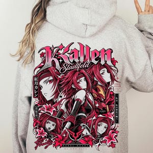 Peut inclure: Un sweat à capuche gris clair avec un grand motif au dos. Le design présente plusieurs illustrations d'un personnage aux cheveux rouges avec le texte Kallen Stadfeld, Code Geass, Guren et des caractères japonais en rose et noir.