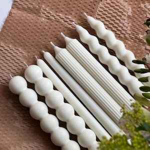 May include: A set of six white candles with different textures. The candles are arranged in a row, with the textured candles in the front and the plain candles in the back.