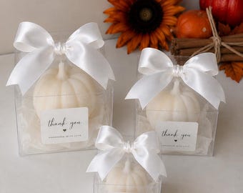 May include: Three clear square gift boxes containing white pumpkin-shaped candles. Each box is topped with a white satin ribbon bow and features a white label with the text thank you and handmade with love printed in black ink.