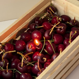 May include: A rectangular light-colored wooden crate filled with numerous dark red cherries. One cherry near the center features a small, lit candle flame emerging from its top, casting a warm glow among the surrounding fruit and thin stems.