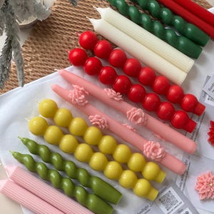 May include: A collection of colorful candles in various designs. Includes red, green, white, pink, and yellow candles. Some are spiral, others have a beaded design, and some are decorated with floral accents. The candles are arranged on a white surface.