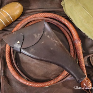 May include: A dark brown leather holster with a flap closure and a single metal snap button, positioned over a coiled braided leather whip. A smooth amber-colored stone with three white stripes and a green canvas bag are also visible.