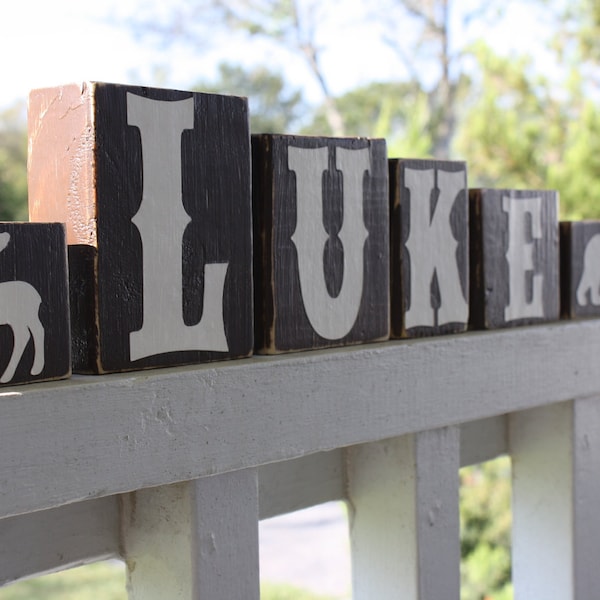 May include: Wooden blocks spelling out the name "LUKE" with a moose and a bear on the ends. The blocks are painted brown with white letters.